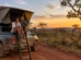 Casal sentado na escada de uma barraca de teto aberta sobre uma caminhonete em estrada de terra no cerrado ao entardecer