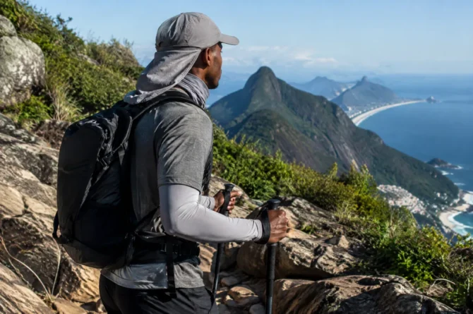Trilheiro de costas usando boné com protetor de nuca e bandana tubular no pescoço durante subida ensolarada