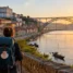 Peregrino com mochila começando o Caminho Português na beira do rio, com casas coloridas e ponte de ferro ao fundo