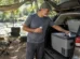 Homem pegando bebida gelada de uma geladeira portátil no porta-malas de um carro estacionado em camping à beira de um rio