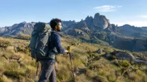 Trekker com mochila cargueira grande atravessando campo de altitude com picos rochosos ao fundo