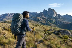 Trekker com mochila cargueira grande atravessando campo de altitude com picos rochosos ao fundo