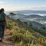 Mulher caminhando em trilha de montanha com mochila de ataque verde, araucárias e vales com neblina ao fundo