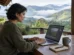 Nômade digital trabalhando num notebook leve numa mesa de varanda de pousada com vista para montanhas
