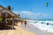 Barracas de praia com cobertura de palha, mar com ondas e pipas de kitesurfe no céu