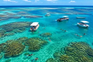 Piscinas naturais azul-turquesa sobre recifes de coral em alto-mar, com barcos e pessoas flutuando
