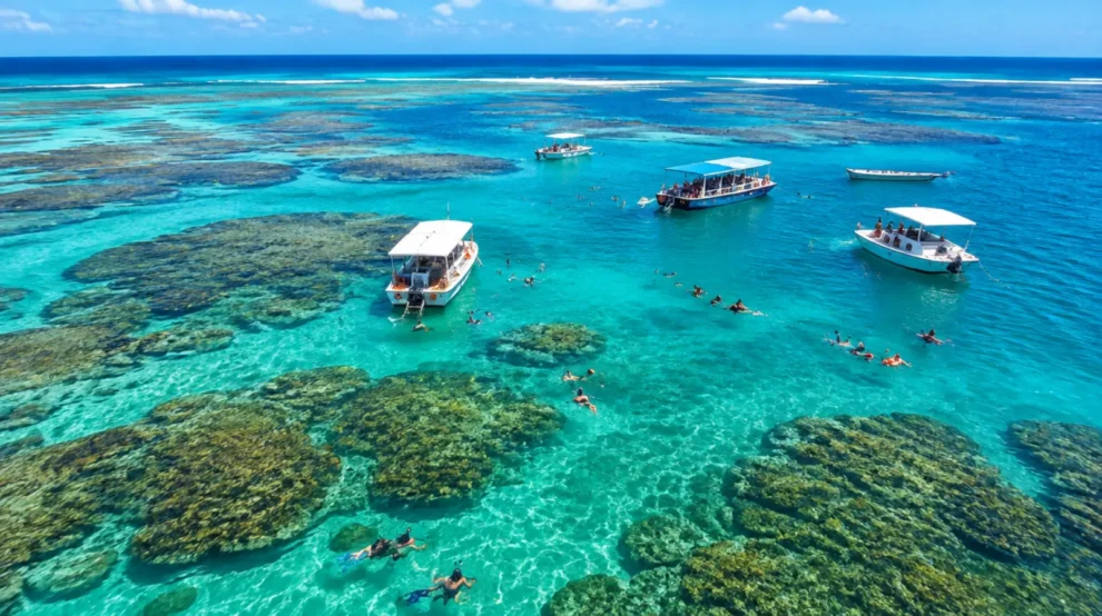 Piscinas naturais azul-turquesa sobre recifes de coral em alto-mar, com barcos e pessoas flutuando