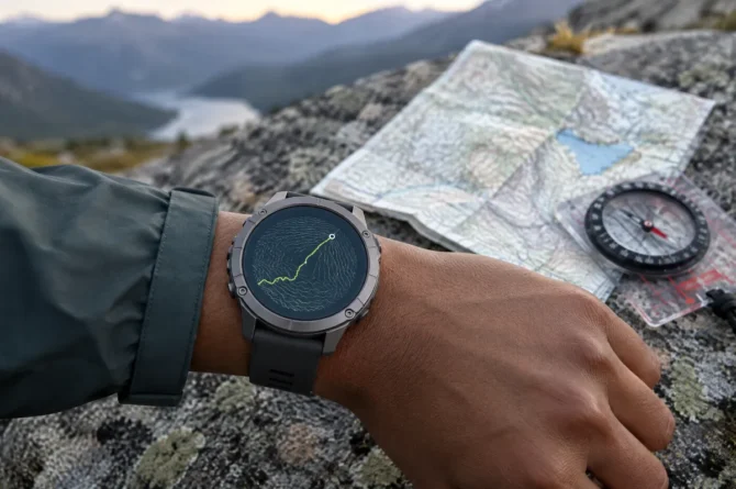 Pulso com relógio GPS robusto mostrando linha de trilha sobre curvas de nível, com mapa de papel dobrado ao fundo