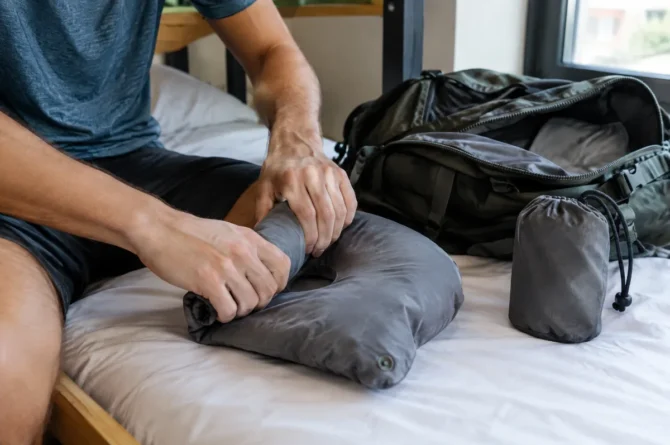 Mãos enrolando um travesseiro inflável vazio ao lado de uma mochila aberta sobre a cama de um hostel
